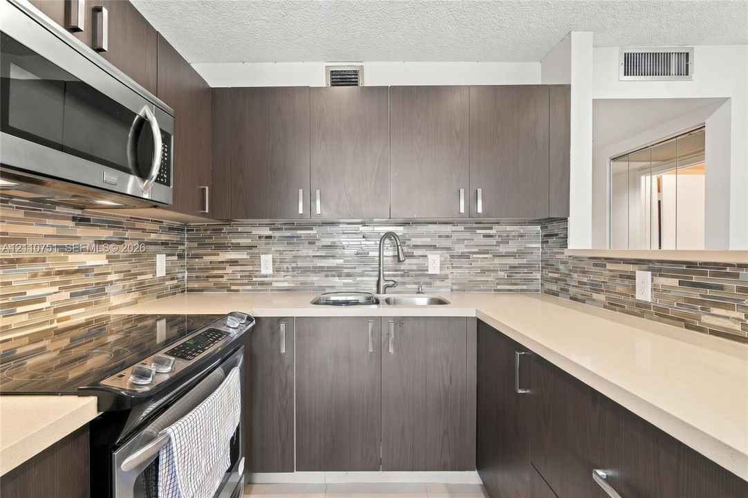 Kitchen featuring dark wood-finish cabinetry, neutral solid-surface countertops, a linear tile backsplash, and stainless steel appliances including a built-in microwave and range