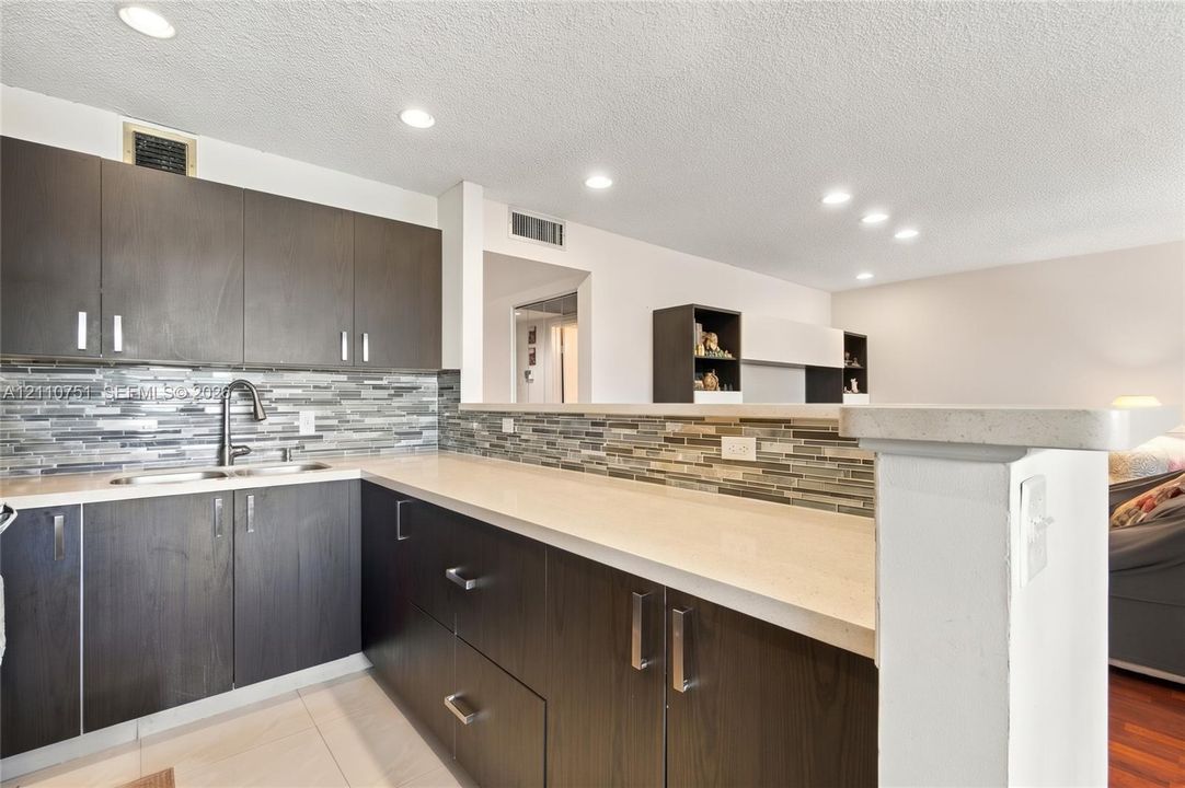 Kitchen featuring dark-toned flat-panel cabinetry, light-colored solid surface countertops, a decorative linear tile backsplash, recessed ceiling lighting, and a high-arc pull-down faucet