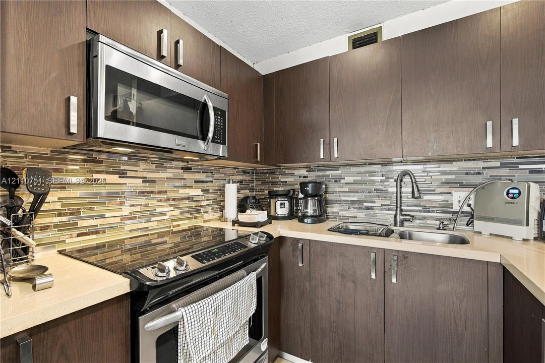 Kitchen featuring dark wood-finish cabinetry, solid-surface countertops, horizontal glass-tiled backsplash, stainless steel microwave, and undermount dual-basin sink with a high-arch faucet