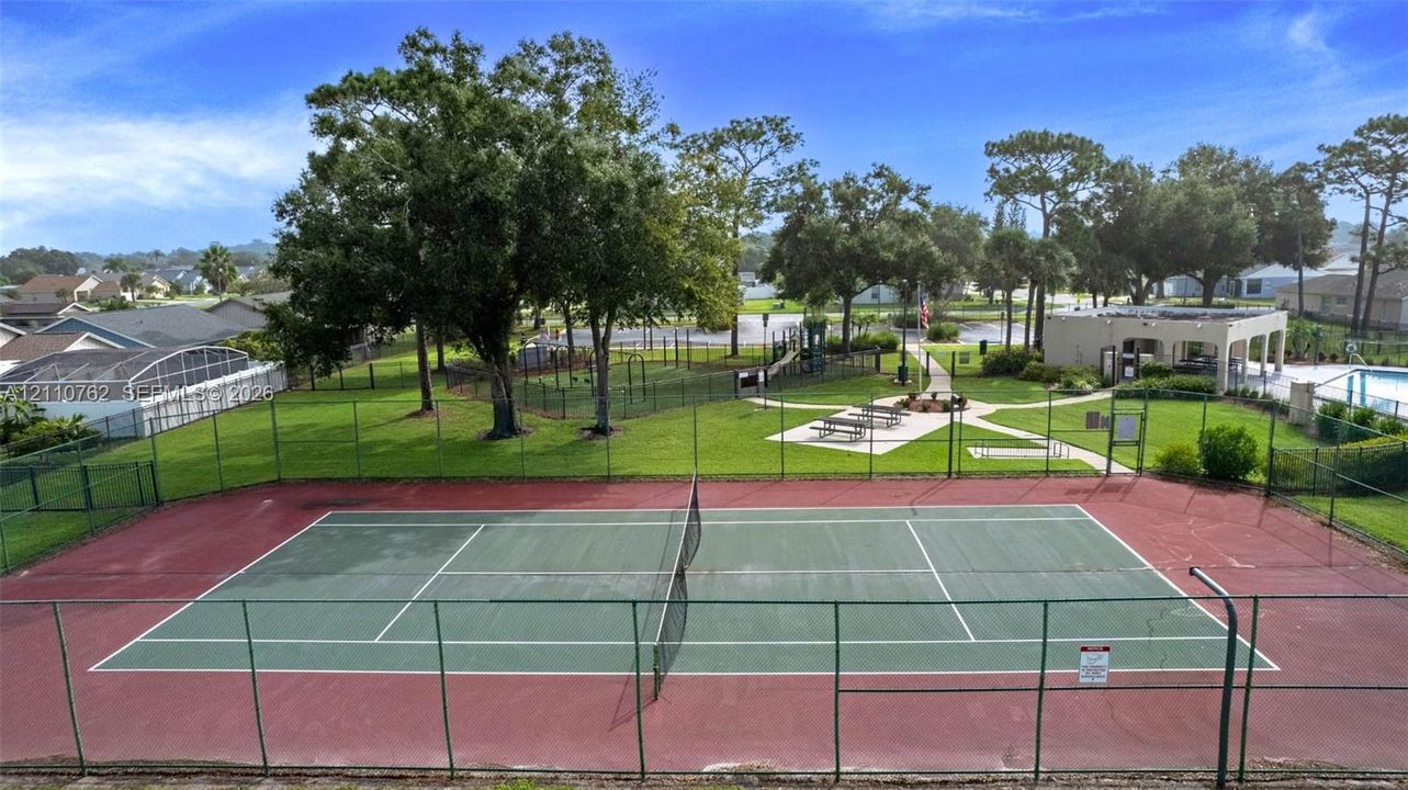 Fenced tennis court featuring a hard surface, complemented by a landscaped common area with mature trees, a swimming pool, and a covered clubhouse