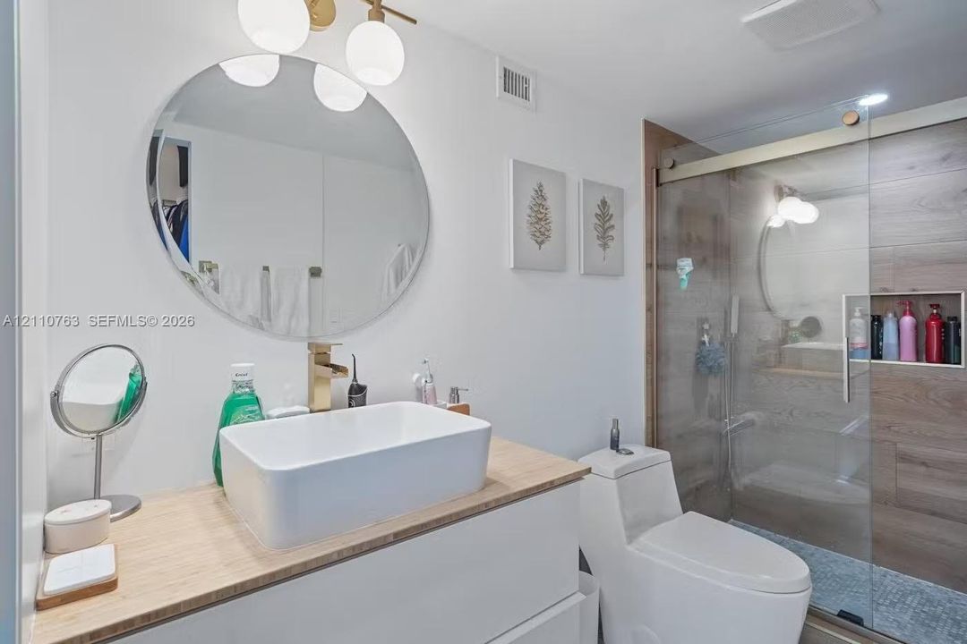 Floating vanity with a light wood-finish countertop, rectangular vessel sink, gold-tone faucet, round wall-mounted mirror, and glass-enclosed shower with wood-finish tile wall and built-in recessed niche