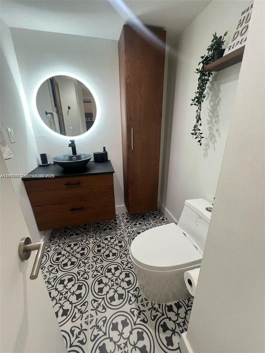 Modern vanity with wood-finish cabinetry, black stone countertop, and circular vessel sink, complemented by a backlit round mirror, floor-to-ceiling wood-finish storage cabinet, and patterned tile flooring