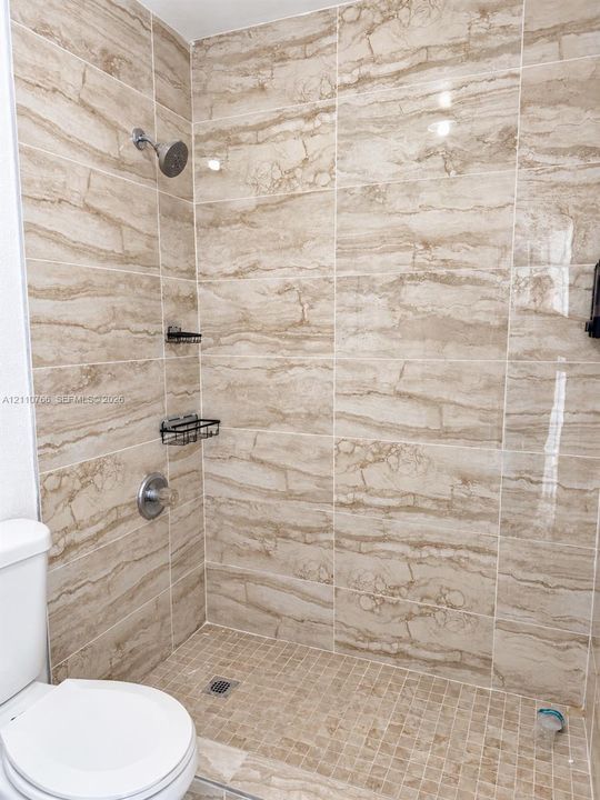Walk-in shower featuring large-format beige tile surround, mosaic tile flooring, built-in metal shelving, and a wall-mounted shower head