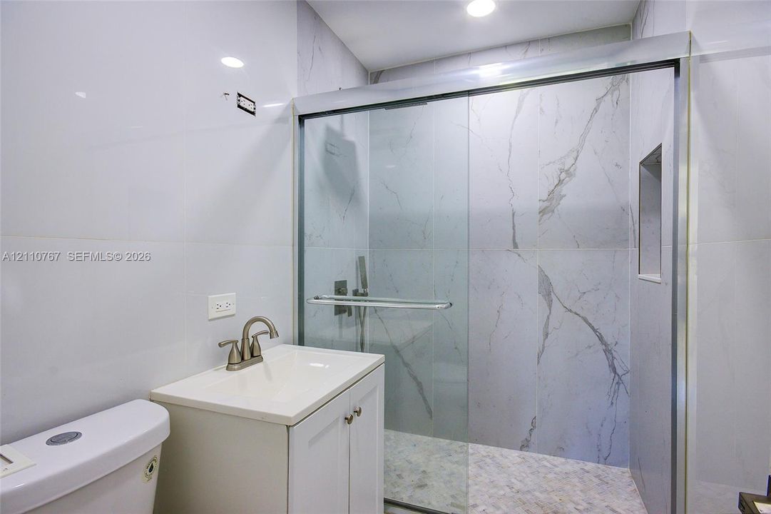 Full-height stone-finish wall tile, frameless glass shower enclosure with sliding door, integrated shower niche, modern vanity cabinet with drop-in sink, and brushed-finish faucet