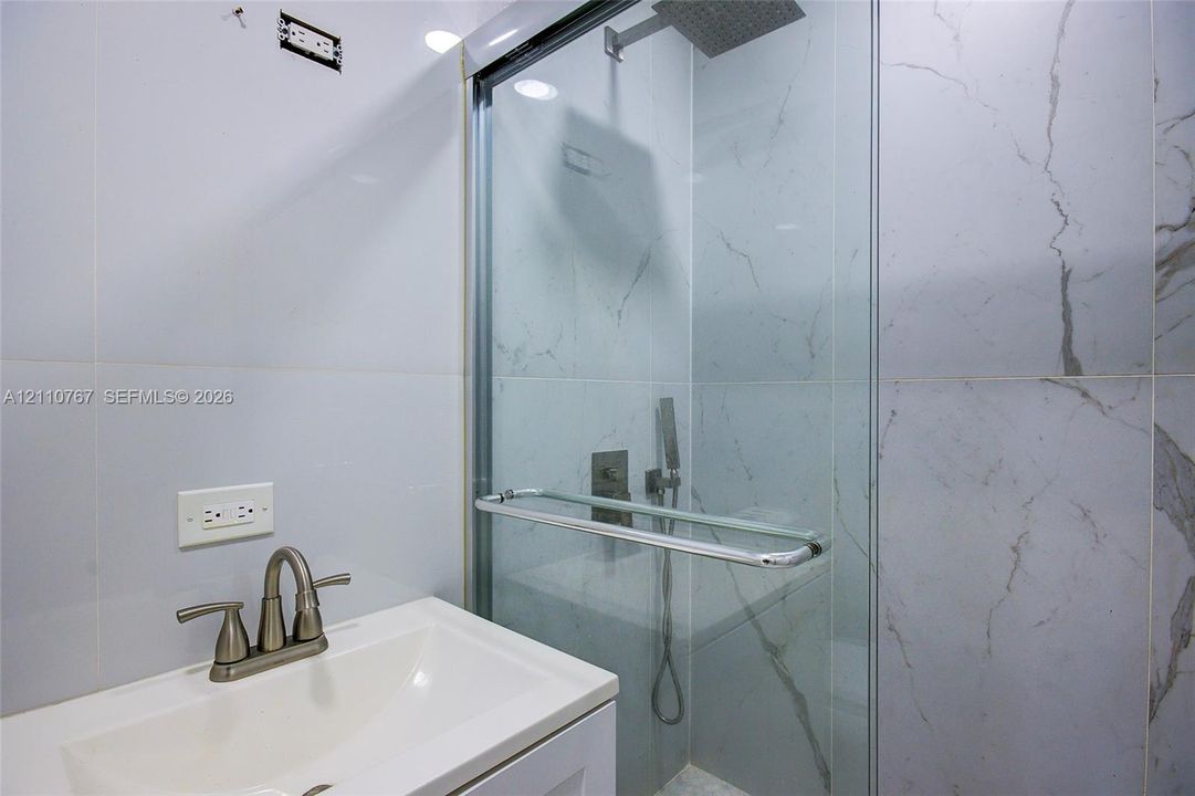Walk-in shower with a glass enclosure, rainfall showerhead, handheld sprayer, and large-format tile surround, featuring a vanity with a white basin and metallic hardware