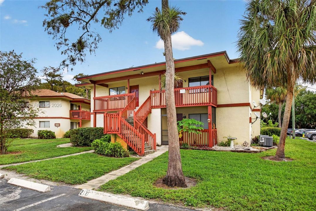 Two-story multi-unit exterior featuring stucco siding, wooden railings, an external staircase, and mature palm trees