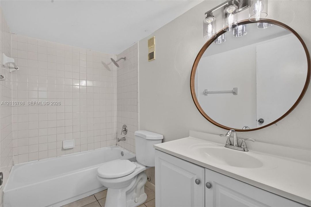 Bathroom featuring a white vanity cabinet with a solid-surface countertop, circular wall-mounted mirror, three-light fixture, and a tiled shower-tub combination