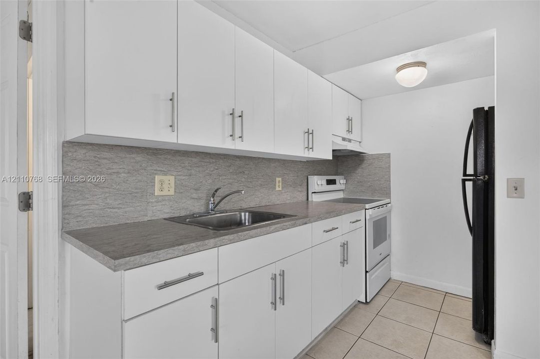 Kitchen featuring white cabinetry with metallic pulls, gray stone-pattern backsplash, neutral countertop, and tile flooring
