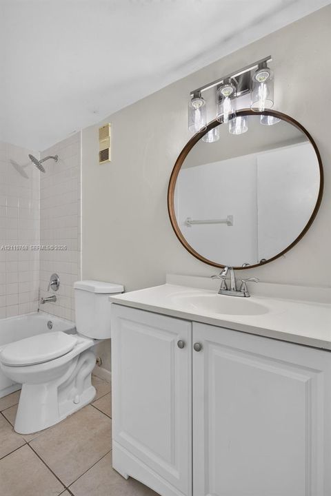 White vanity with a solid-surface countertop and integrated sink, round mirror with a bronze-toned frame, three-light fixture with glass shades, and tiled tub surround
