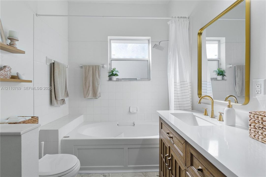 Bathroom featuring a wood-finish vanity with a stone-surface countertop, gold-tone faucet, rectangular wall-mounted mirror with a metallic frame, and a built-in soaking tub with a tiled surround