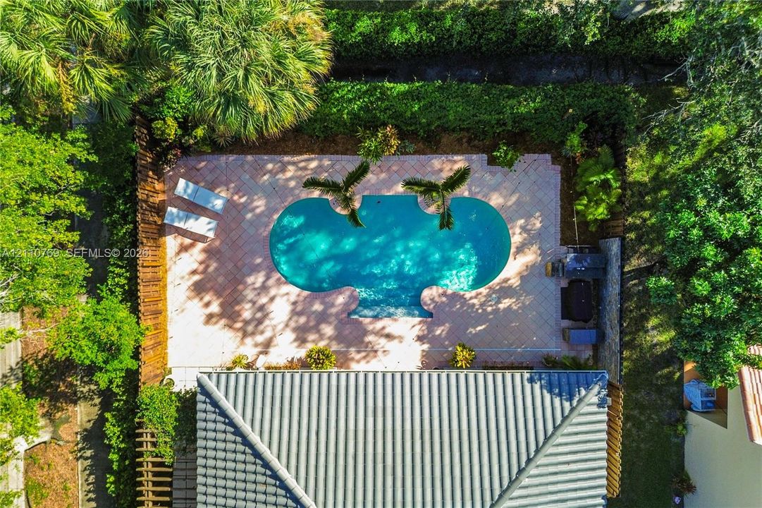 Custom-shaped swimming pool featuring a spacious paver patio, encased within a private, fenced perimeter surrounded by mature tropical landscaping