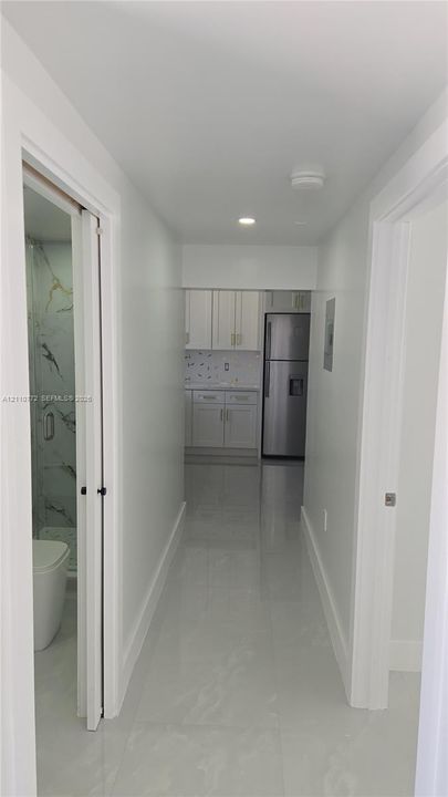 Hallway featuring light-toned flooring, recessed lighting, and access to a bathroom with a glass-enclosed shower and a kitchen equipped with white cabinetry and a stainless steel refrigerator