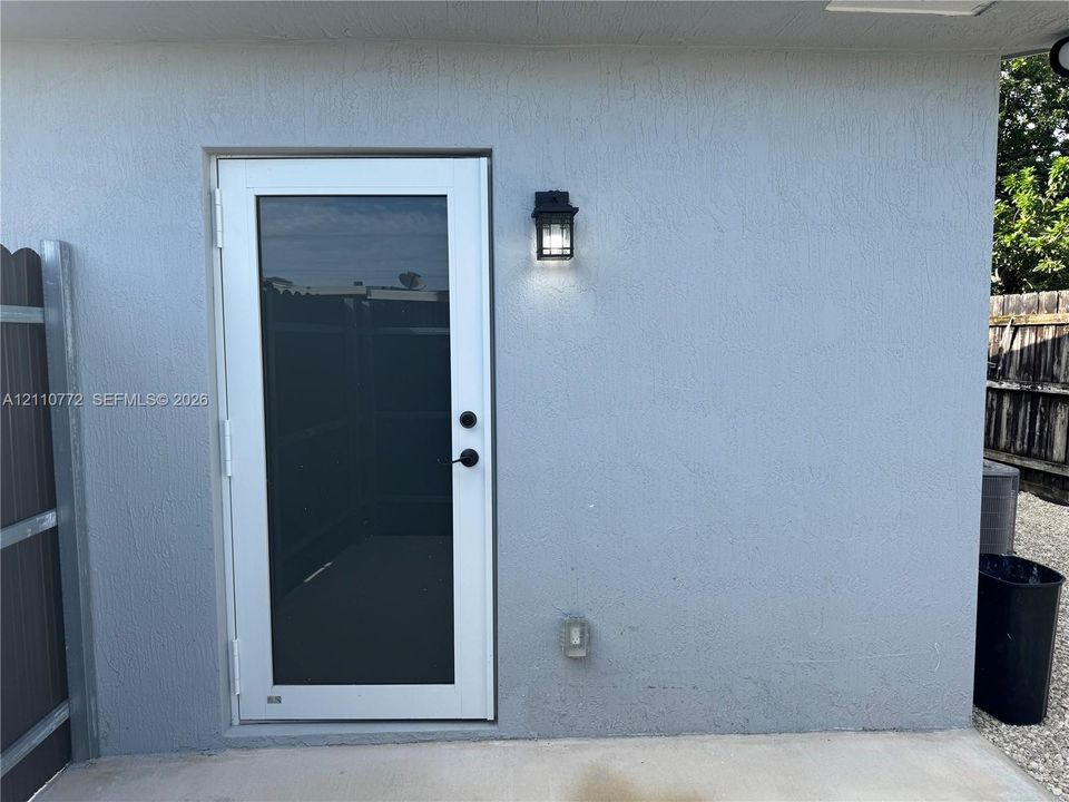Exterior door featuring a white frame with a full-length glass pane, matte black hardware, and an adjacent wall-mounted lantern-style exterior light fixture against a stucco finish