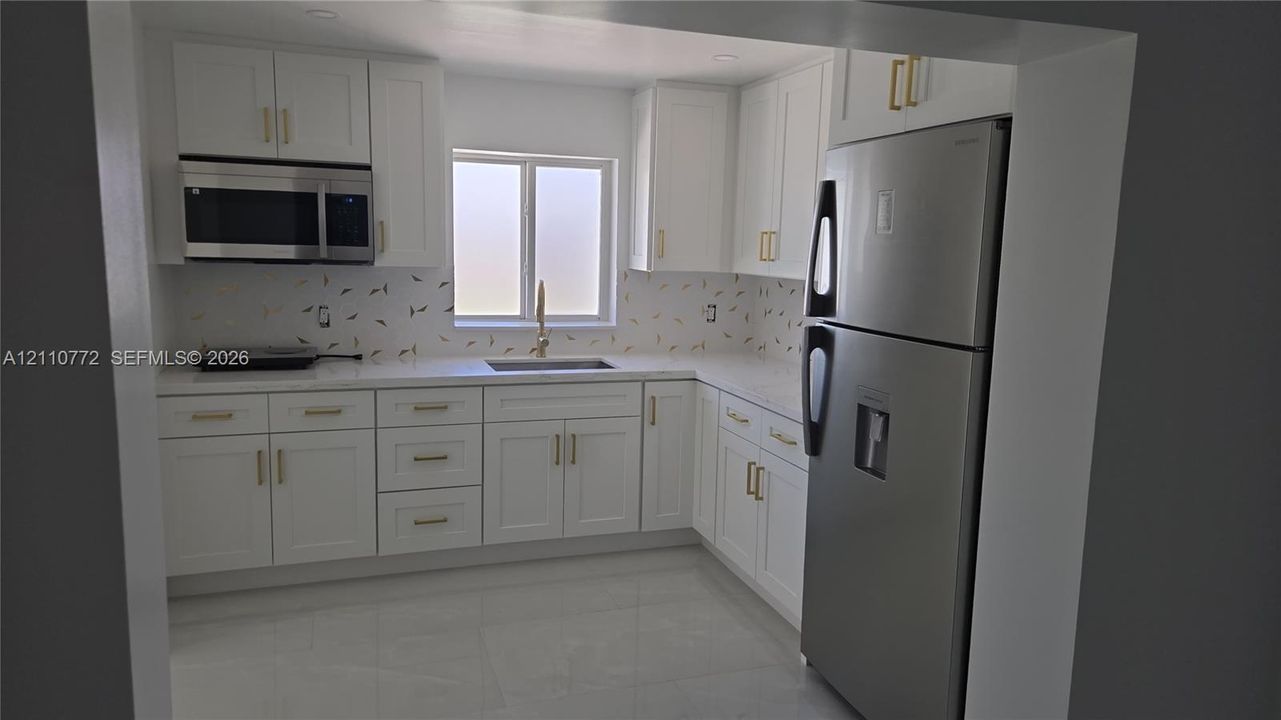 Kitchen featuring white shaker-style cabinetry, gold-toned hardware, stone-finish countertops, stainless steel appliances, and a patterned tile backsplash