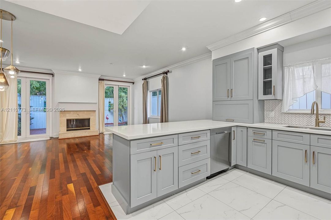 Kitchen featuring gray shaker-style cabinetry, quartz countertops, a herringbone-patterned tile backsplash, wood-finish flooring, and a stone-surround fireplace