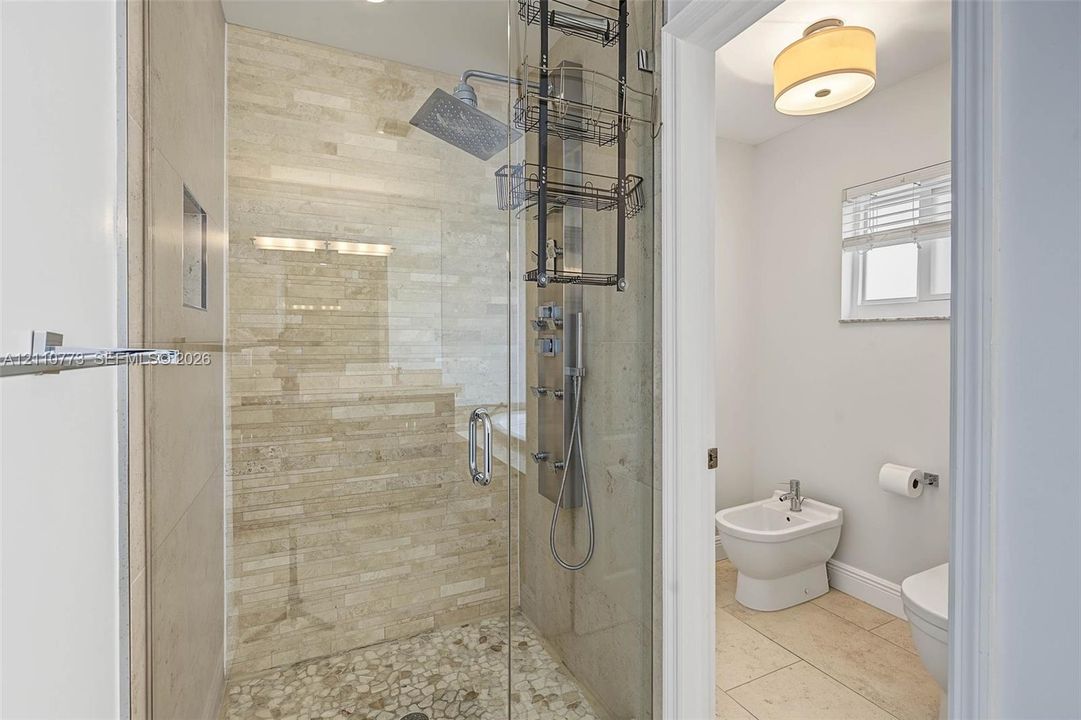 Walk-in shower featuring multi-head shower system, stone-tiled walls, and pebble-style floor, paired with a bidet and stone-tile flooring