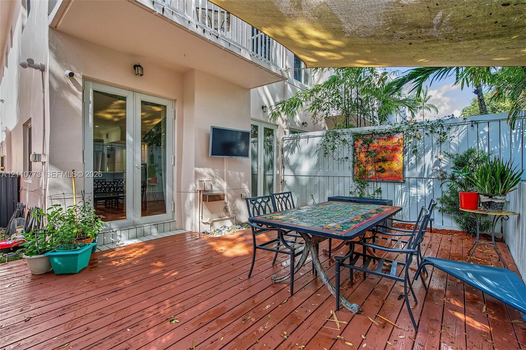 Exterior wood-finish deck with a textile shade sail, double glass-panel doors, and white privacy fencing