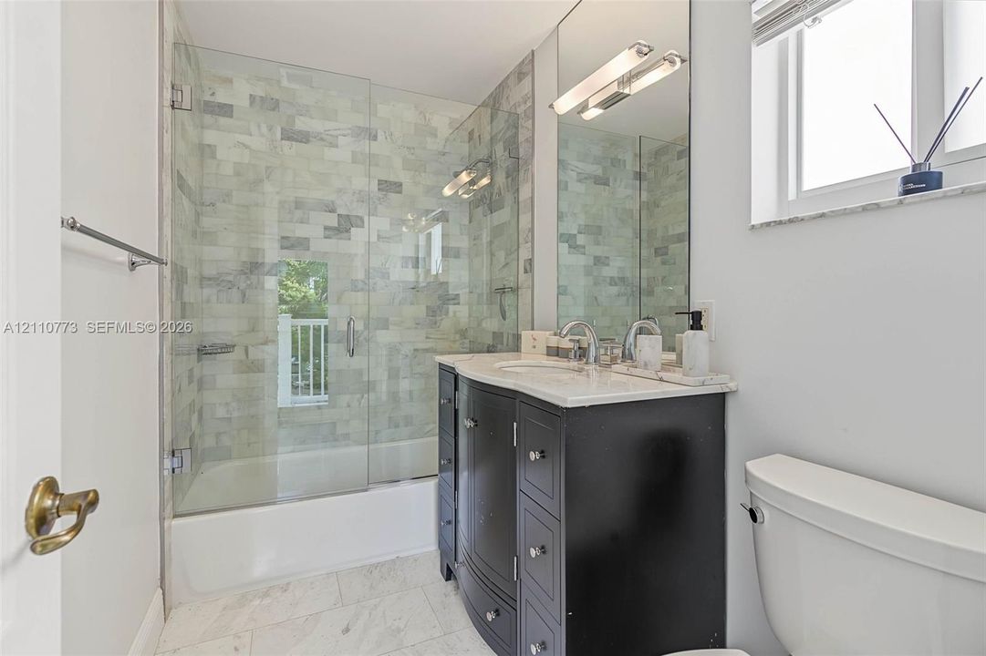 Curved vanity cabinet featuring a stone-surface countertop, glass-enclosed tub and shower combination with stone tile surround, full-width mirror, and dual-light wall-mounted fixture