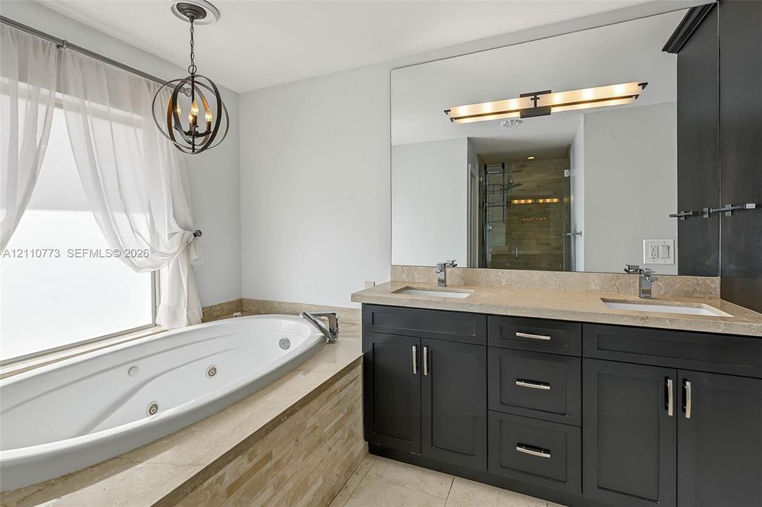 Dark wood cabinetry with stone countertop, dual undermount sinks, jetted soaking tub with stone surround, and an orb-style pendant light fixture