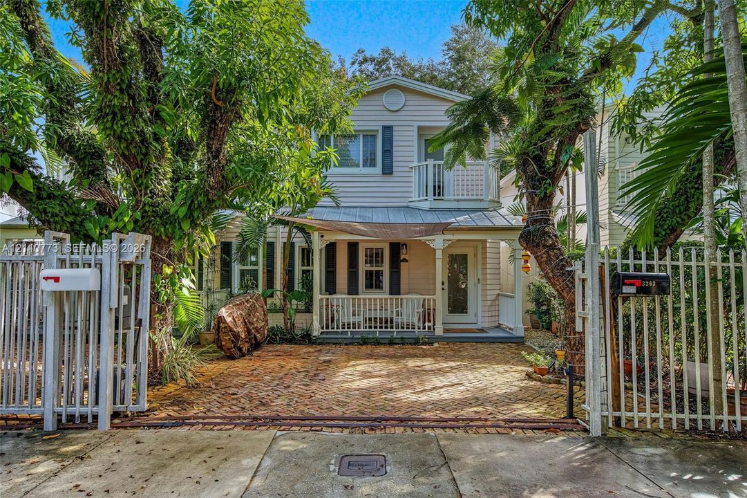 Two-story residence featuring light-colored horizontal siding, a covered front porch with white railing, a metal roof accent, and a fenced-in brick-paved courtyard