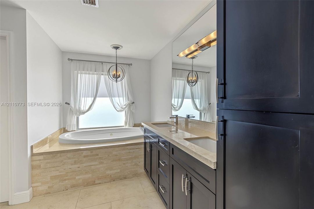 Double vanity with dark cabinetry and stone countertop, large wall-mounted mirror, soaking tub with tile surround, and spherical pendant lighting