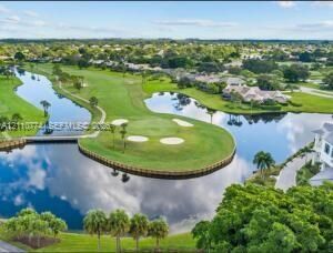 Aerial perspective of a golf course featuring manicured greens, sand bunkers, expansive water elements, and a paved pedestrian bridge