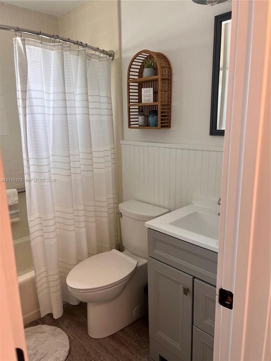 Bathroom featuring a grey vanity with a rectangular sink, beadboard wainscoting, a bathtub with tile surround, and wood-finish flooring