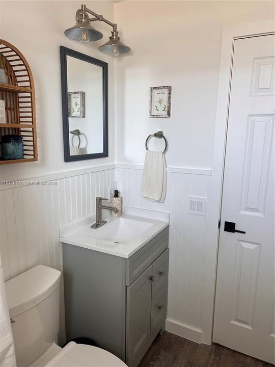 Gray vanity with a white countertop and basin, white beadboard wainscoting, dual-light metal vanity fixture, framed wall mirror, and six-panel interior door with matte black hardware