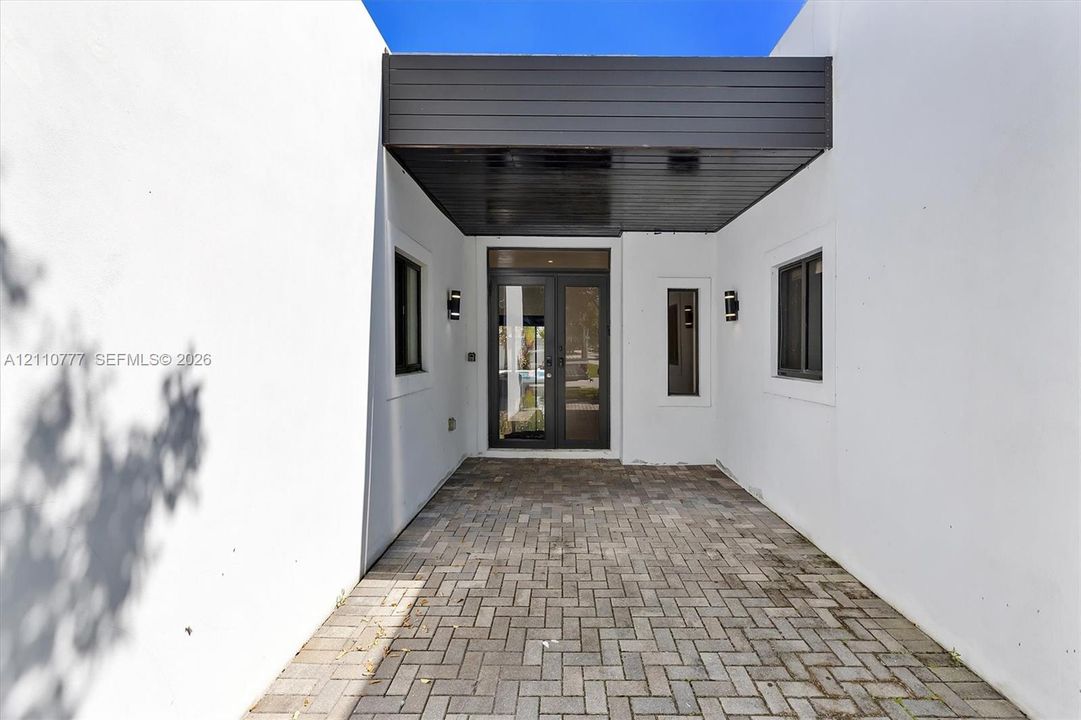 Covered entryway featuring a herringbone-patterned paver floor, minimalist smooth-stucco exterior walls, dark wood-finish ceiling paneling, glass-paneled double doors, and wall-mounted exterior sconces