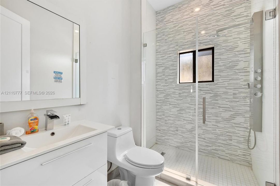 Modern bathroom featuring a white vanity with an integrated countertop, wall-mounted mirror, walk-in shower with a glass enclosure, patterned tile shower wall, and an integrated shower panel system