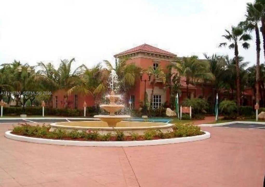 Tiered stone fountain centered in a circular driveway, framed by mature palms, low-profile shrubbery, and Mediterranean-inspired architecture featuring a tile roof