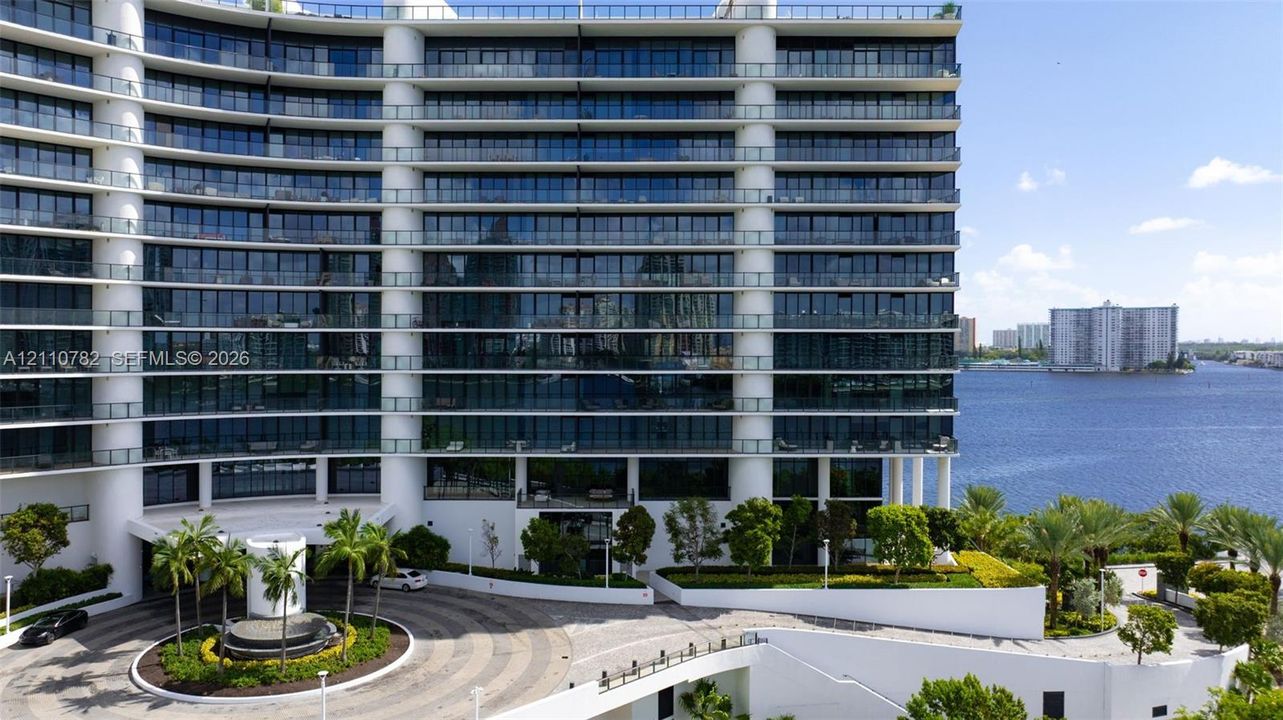 Mid-rise architectural exterior featuring tiered glass balconies, floor-to-ceiling windows, white structural columns, a circular paved motor court, and waterfront landscaping