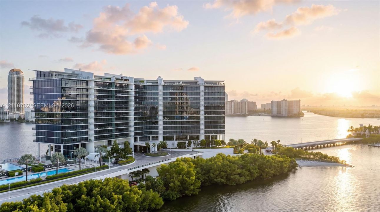 High-rise residential building featuring floor-to-ceiling glass paneling, wrap-around balconies, a porte-cochère entryway, and an outdoor swimming pool, situated on a waterfront lot with dense perimeter foliage
