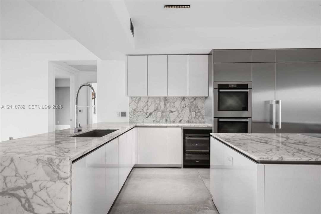 Kitchen featuring white flat-panel cabinetry, stone countertops with waterfall edge, full-height stone backsplash, stainless steel double wall ovens, and an under-counter beverage cooler