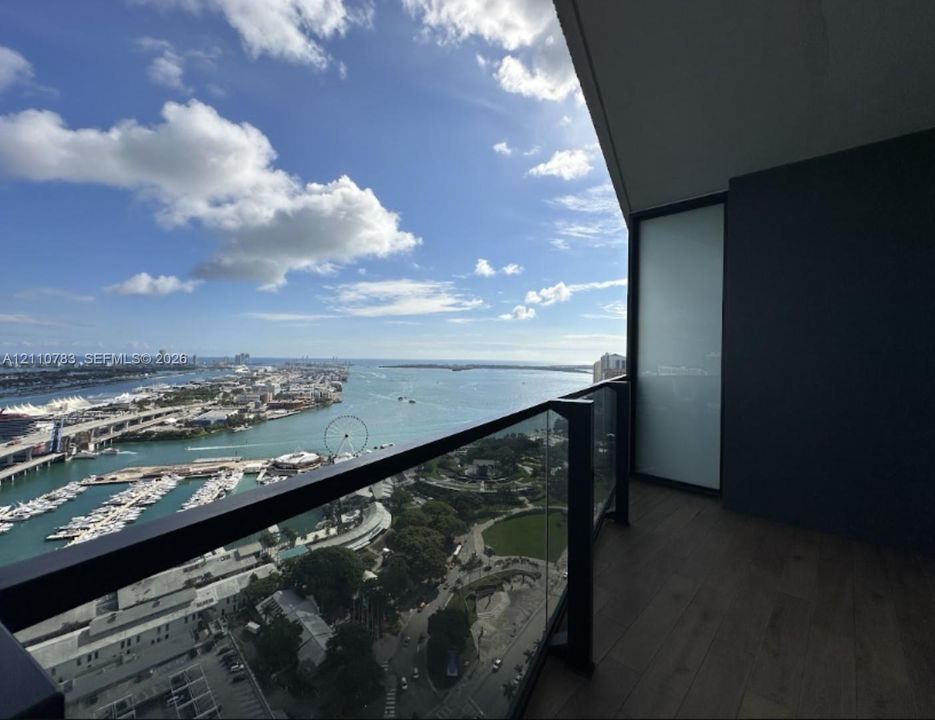 Covered balcony featuring wood-finish flooring, a sleek glass railing with black framing, and expansive water and urban scenery