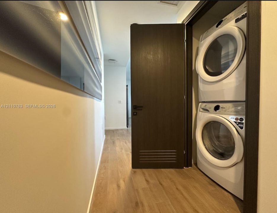 Laundry closet featuring a stacked front-loading washer and dryer, finished with a dark wood-tone door and coordinating wood-finish flooring