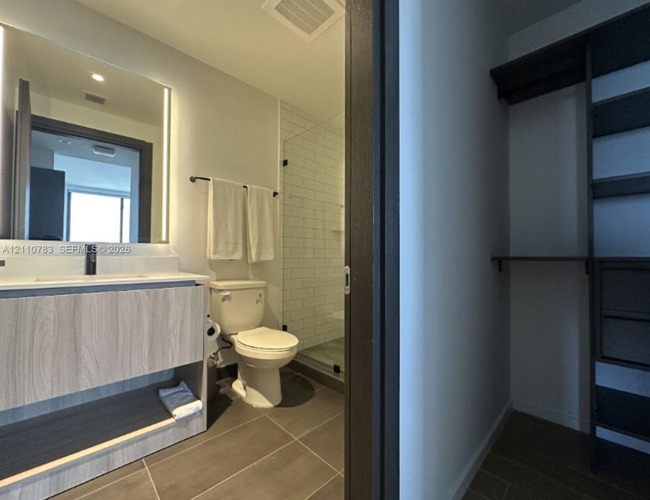 Bathroom featuring a wood-finish vanity with a light countertop, backlit mirror, matte black fixtures, tile flooring, and a glass-enclosed shower with subway tile surround, accompanied by a custom built-in closet system