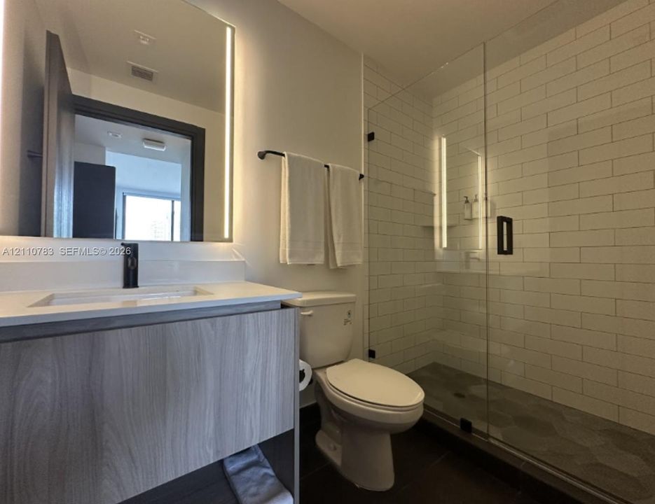 Modern vanity with a light wood-finish cabinet and integrated sink, frameless glass shower enclosure, subway tile shower surround, and LED-integrated wall mirror