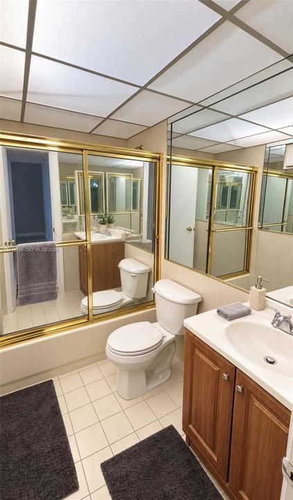Bathroom featuring a wood-finish vanity with a drop-in sink, ceramic tile flooring, a shower-tub combination with brass-tone sliding doors, wall-to-wall mirror, and a grid-panel drop ceiling
