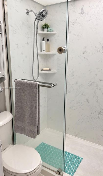 Walk-in shower enclosure featuring light-toned stone-finish wall panels, a handheld showerhead, built-in corner shelving, and a frameless glass door with a polished metal handle