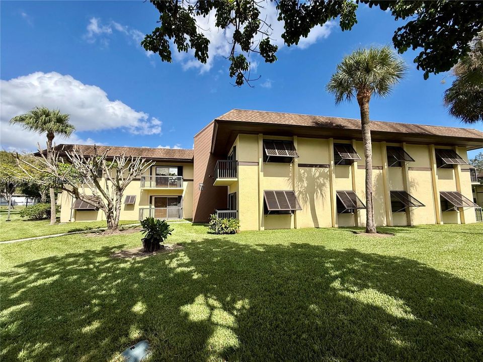 Multi-story exterior featuring stucco siding, metal window awnings, balcony access, and surrounding landscaped grounds with mature palm trees