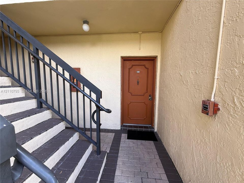 Exterior entry featuring a paneled wood-finish door, paver tile flooring, and metal railing