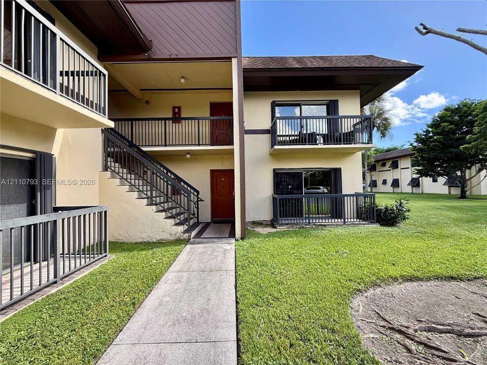 Exterior view featuring multi-level balconies with dark metal railings, concrete stairs, and paved walkway set within a manicured lawn