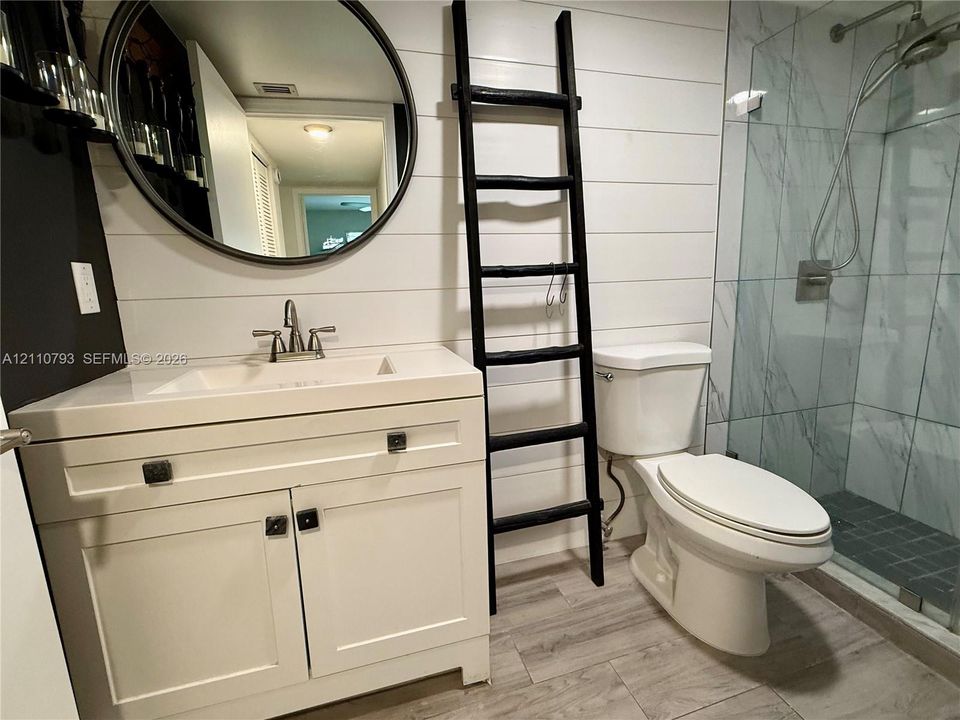 Bathroom featuring white horizontal shiplap wall paneling, wood-finish flooring, a white vanity with an integrated basin, a frameless glass-enclosed shower with tiled walls, and a round wall-mounted mirror