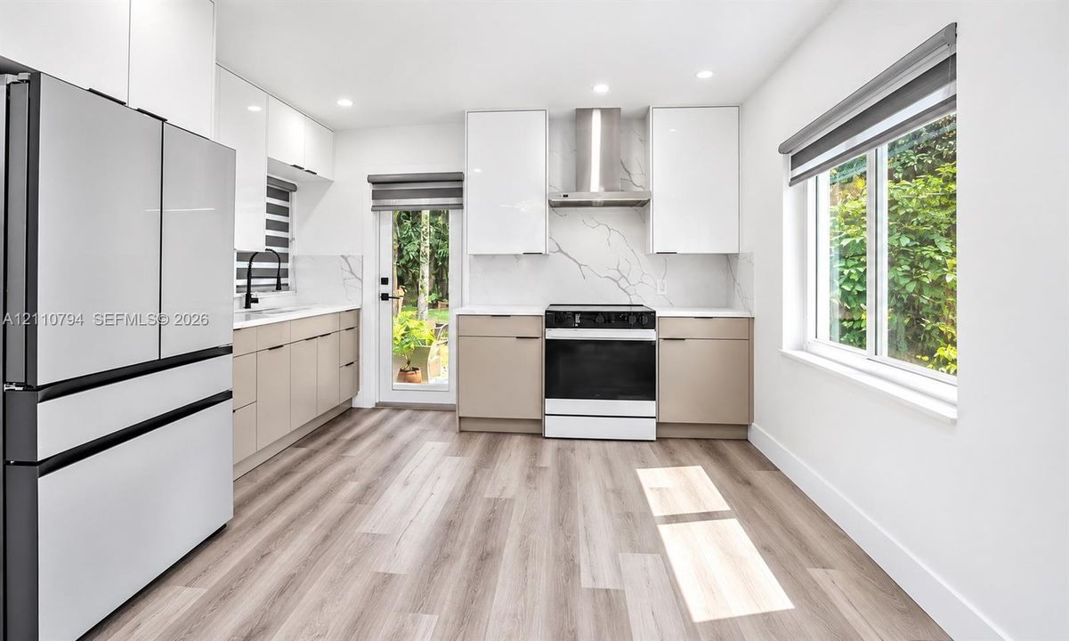 Kitchen featuring high-gloss cabinetry, stone-patterned backsplash, stainless steel range hood, wood-finish flooring, and a glass-paneled exterior door