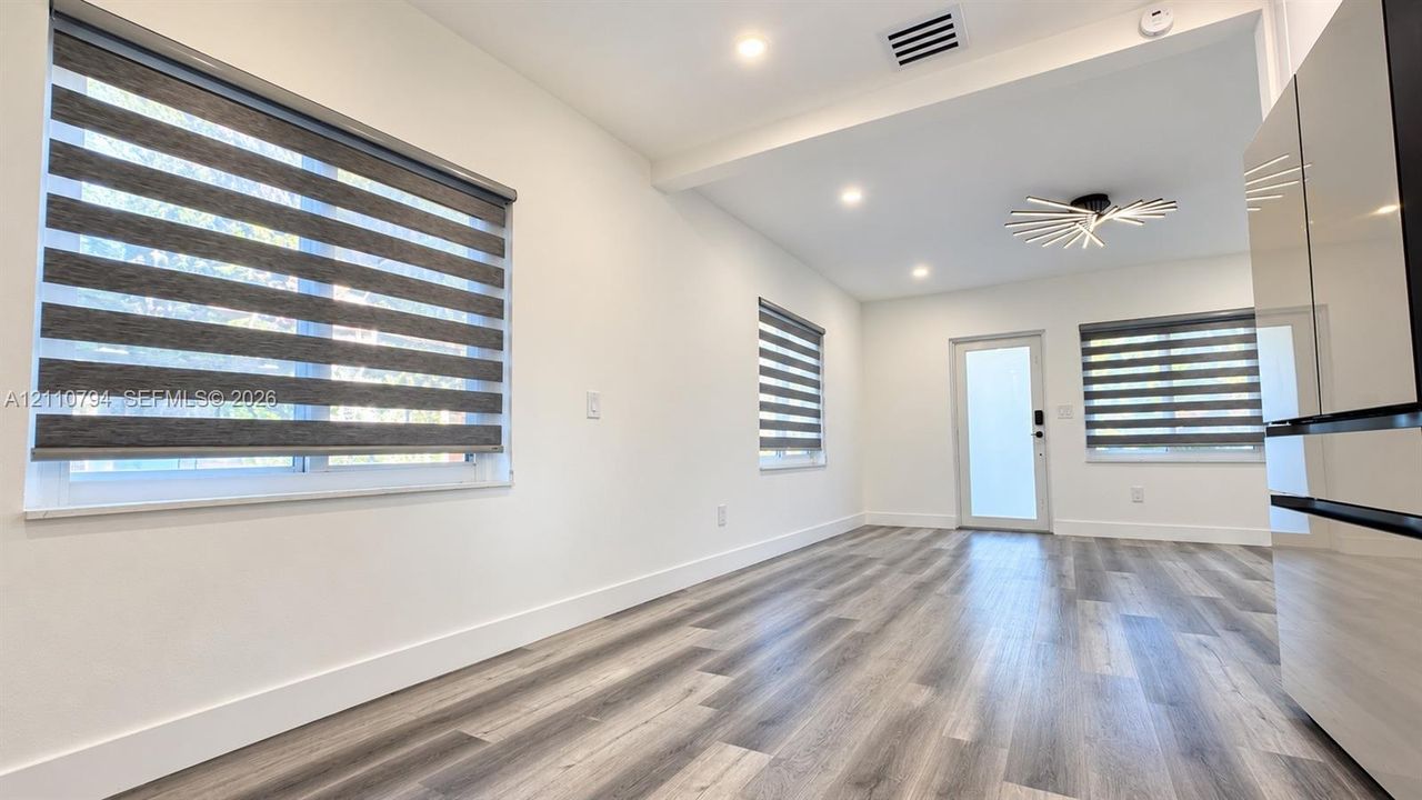 Wood-finish flooring, multi-layered roller shades, recessed ceiling lighting, contemporary geometric overhead fixture, and full-glass exterior entry door