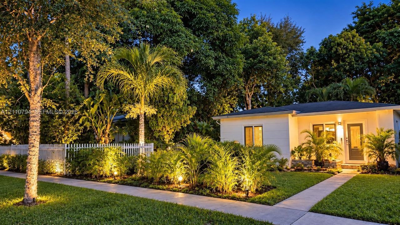 Single-story residence featuring light-colored horizontal siding, a white picket fence, a concrete walkway, and established landscaping with palm trees