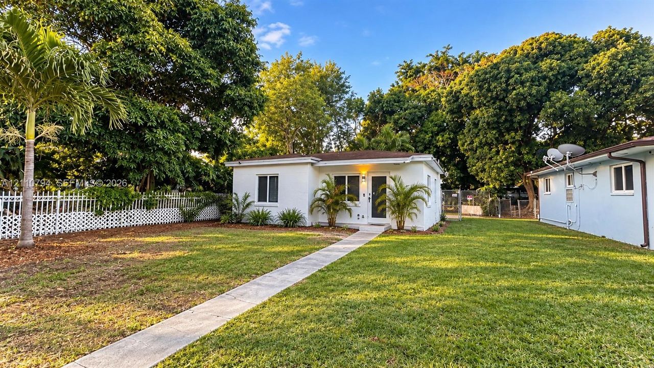 Single-story exterior featuring white stucco siding, a concrete pathway, mature trees, and white lattice fencing
