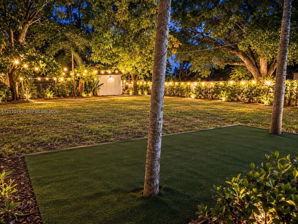 Expansive backyard featuring synthetic turf, mature trees, a detached storage structure, perimeter fencing, and integrated landscape lighting