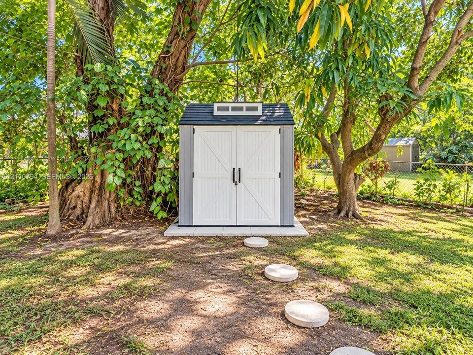 Exterior storage shed featuring dual white doors, a gabled roof with a transom window, and a stone paver foundation set within a landscaped yard